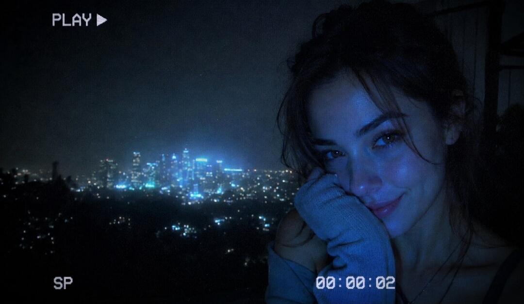 PLAY ▶ SP 00:00:02. A young woman smiles at the camera with a city skyline at night in the background, captured with a retro VHS aesthetic.