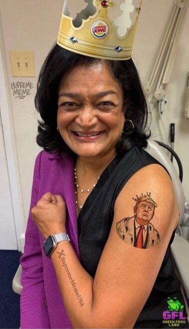 Cartoon tattoo of Donald Trump wearing a crown on the upper arm. Vertical text along the forearm (not fully legible). Wall graffiti reads 'SUPREME MEME'.