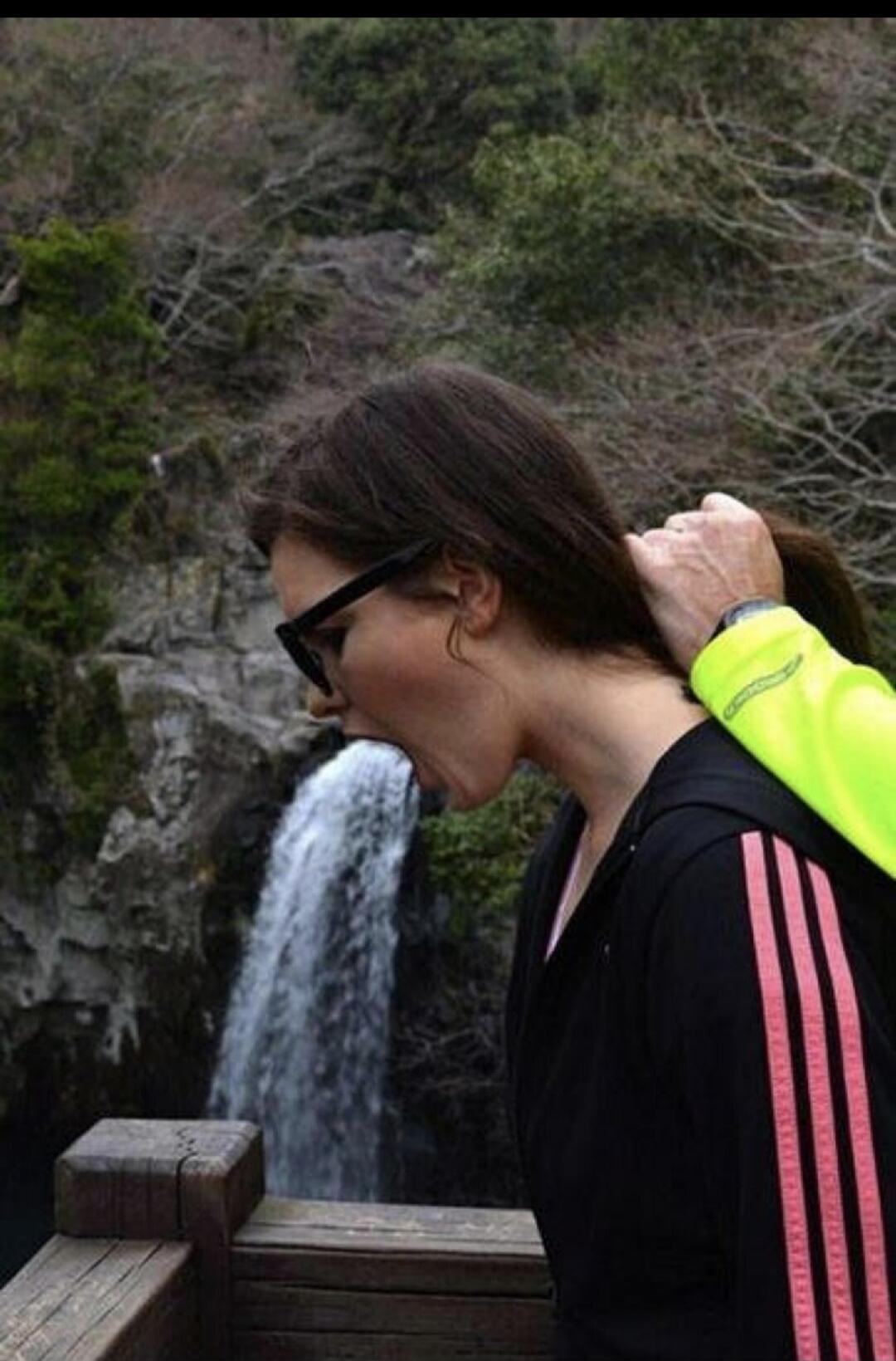 The image shows a woman with her mouth open, appearing to be spitting water from a waterfall. A hand is pulling her hair from behind.