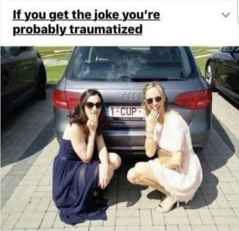 If you get the joke you're probably traumatized. Two women are squatting behind a car with a license plate that reads '1-CUP'. Both women are covering their mouths, appearing to be either shocked or laughing.