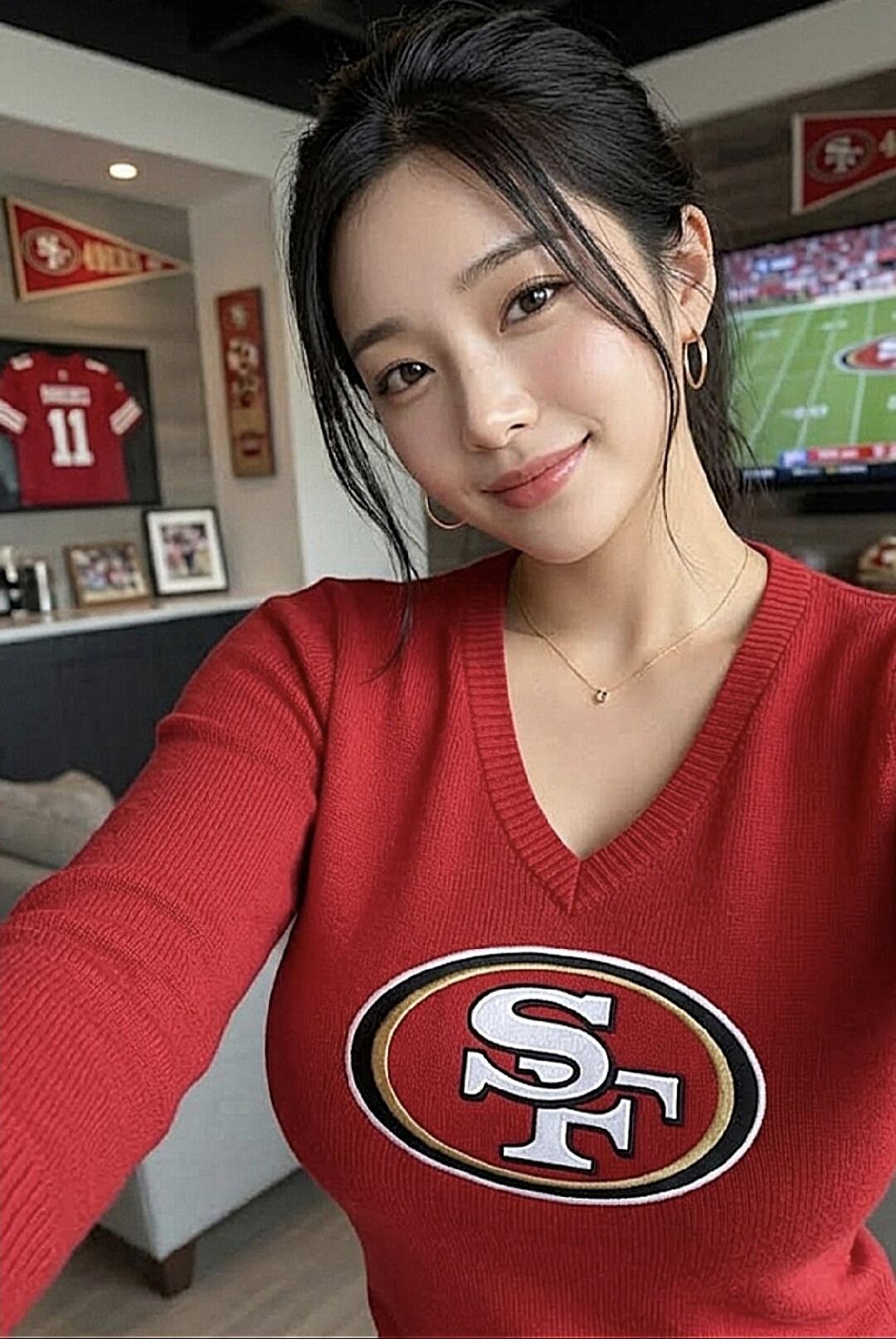 A young woman in a red sweater with the San Francisco 49ers logo, smiling at the camera. In the background, there's a football jersey with number 11, a team pennant, and a TV screen showing a football game.