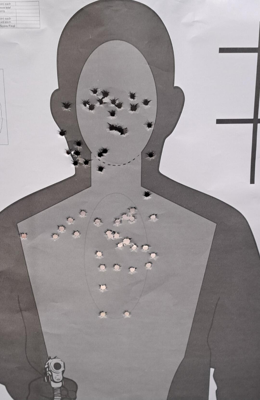 A paper shooting target in the shape of a human silhouette, heavily riddled with bullet holes in the head and chest area. A hand holding a pistol is visible at the bottom.