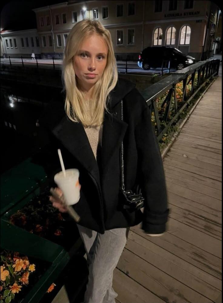 A young blonde woman stands on a wooden bridge at night, holding a white cup with a straw. She is wearing a black coat and light-colored pants. In the background, there are illuminated buildings, one of which has 'KATHEDRAL KYRKAN' written on it. A canal runs alongside the bridge, and flowers are visible in planters along the railing.