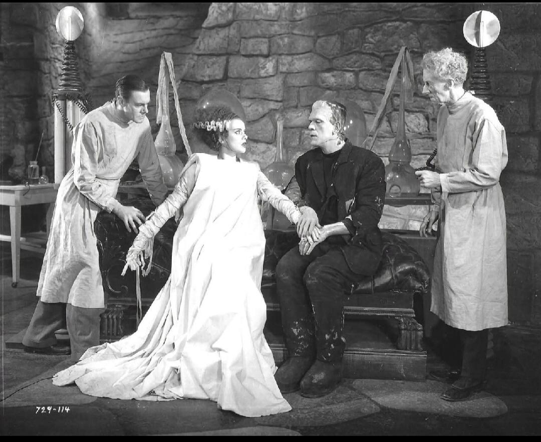A black-and-white scene featuring a woman in a long white dress seated between two men, flanked by two attendants in lab coats, in a stone-walled set with medieval-style stair rails.