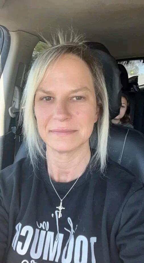 A woman with blonde hair is taking a selfie in a car. A child is partially visible in the back seat. The woman is wearing a black sweatshirt with a cross necklace.