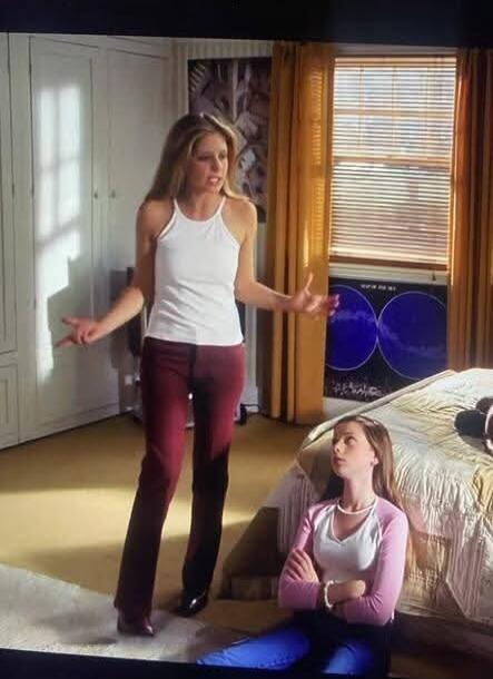 A woman stands in a bedroom, gesturing with her hands as if explaining or arguing, while another woman sits on the floor, looking up at her.