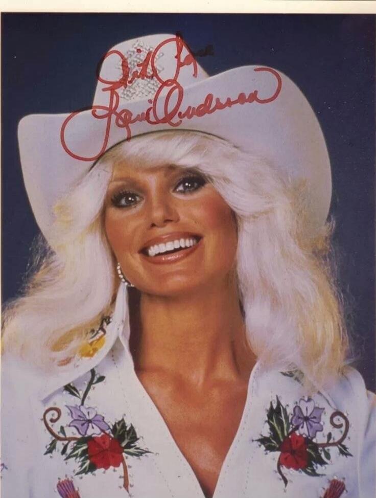 The image shows a close-up of a woman wearing a white cowboy hat and a white shirt with floral embroidery. There is a red signature on the hat. The woman has blonde hair, heavy eye makeup, and a bright smile. The overall aesthetic suggests a vintage country music or rodeo theme.