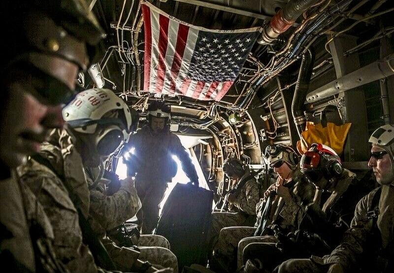 Soldiers or Marines in military gear are seated inside a transport aircraft or helicopter. An American flag is prominently displayed, hanging from the ceiling. Text visible on the flag includes 'BEAR' and '6-17-15'. One helmet has '161 08' written on it. A bright light source is visible in the background.