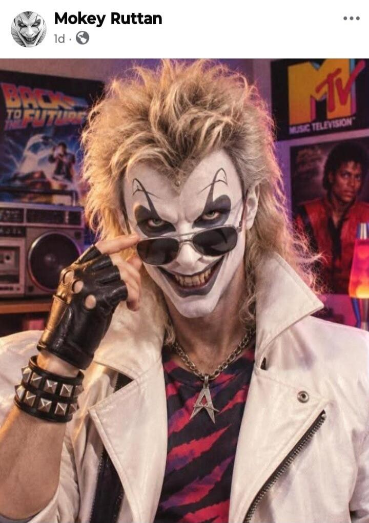 Mokey Ruttan 1d. A person dressed as a clown or joker character, with white face paint, black eye makeup, spiky blonde hair, sunglasses, a white leather jacket, a striped shirt, fingerless gloves, and a studded wristband. In the background, there are posters for 'Back to the Future' and 'MTV Music Television' with Michael Jackson, along with a boom