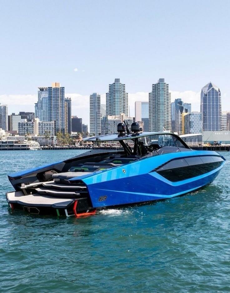 A luxurious blue and black yacht, possibly a Lamborghini Tecnomar 63, on the water with a city skyline in the background under a clear blue sky. The number '63' is visible on the stern.