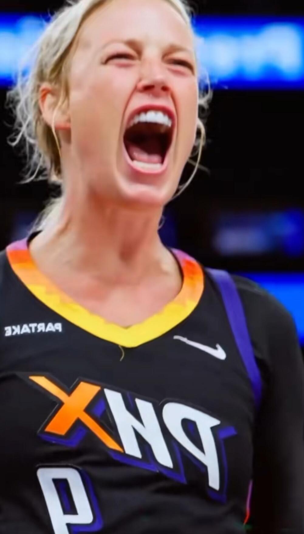 A blonde woman in a black and orange Phoenix Mercury basketball jersey is screaming intensely, showing strong emotion during a game. The jersey has 'PHX' and a Nike logo. Reversed text 'ЗАТАЯЧ' (likely 'PARTNERS') is also visible.