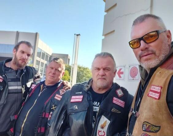 Four men, members of the Hells Angels Motorcycle Club, are posing for a photo outdoors. They are wearing leather vests with various patches, including 'Hells Angels MC', 'Russia', 'Moscow', and 'Support 81'. In the background, there's a building and a wall with a red arrow and a spiral symbol.