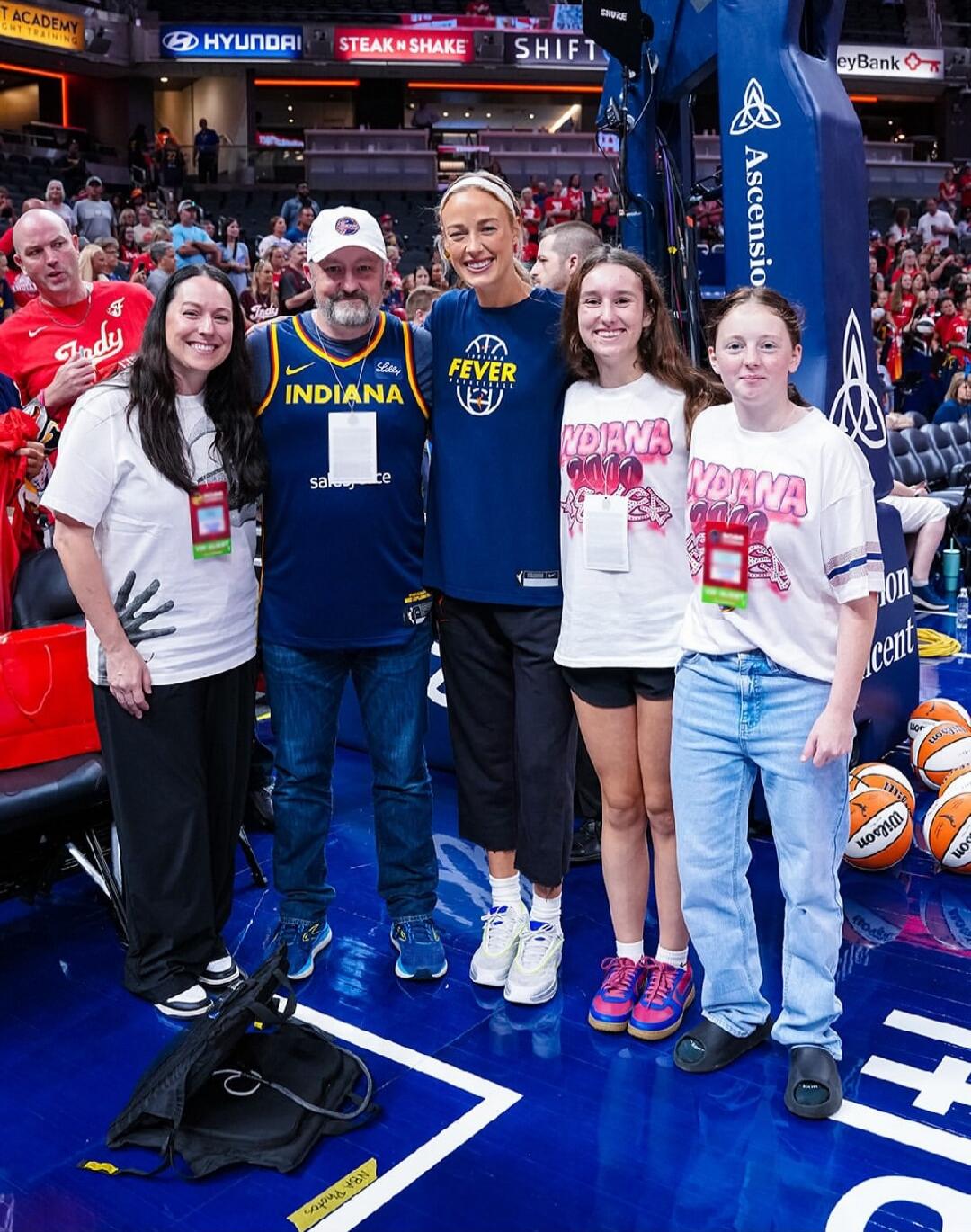 A group of people, including a woman in an Indiana Fever jersey and a man in an Indiana Pacers jersey, posing for a photo on a basketball court. Two girls are wearing 'Indiana' t-shirts. The background shows a basketball arena with spectators and sponsor signs like Hyundai, Steak 'n Shake, Shift, Shure, and Ascension. Basketballs are visible on the