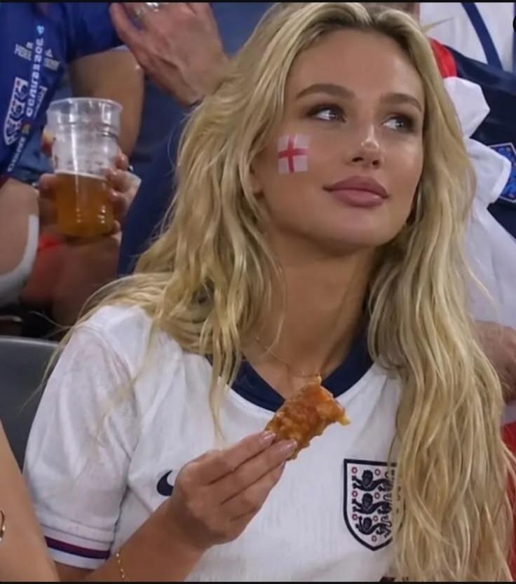 A blonde woman wearing an England football jersey with an England flag painted on her cheek, holding a piece of fried food. She appears to be watching a game.