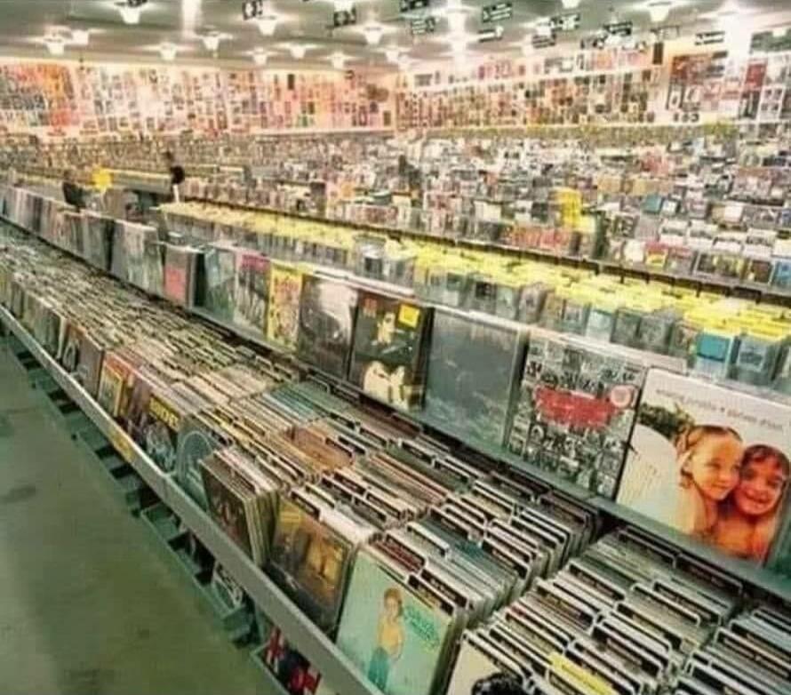 A large record store with long rows of shelves stacked with vinyl records. Album covers are visible from various angles, creating a colorful mosaic of discs.