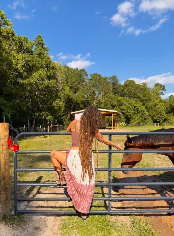 A person with long dreadlocks and tattoos, wearing a crop top and a long skirt, is climbing over a fence to interact with a horse in a sunny, green field.