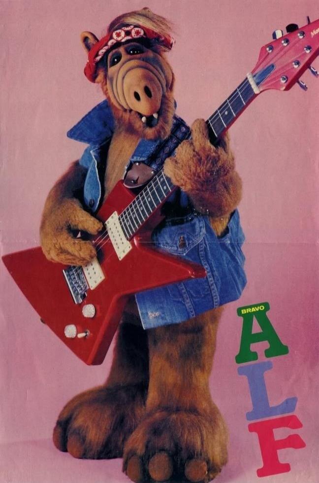 BRAVO ALF. The character ALF, an alien from the planet Melmac, is depicted playing a red electric guitar. He is wearing a denim vest, denim shorts, and a red bandana on his head. The background is pink.