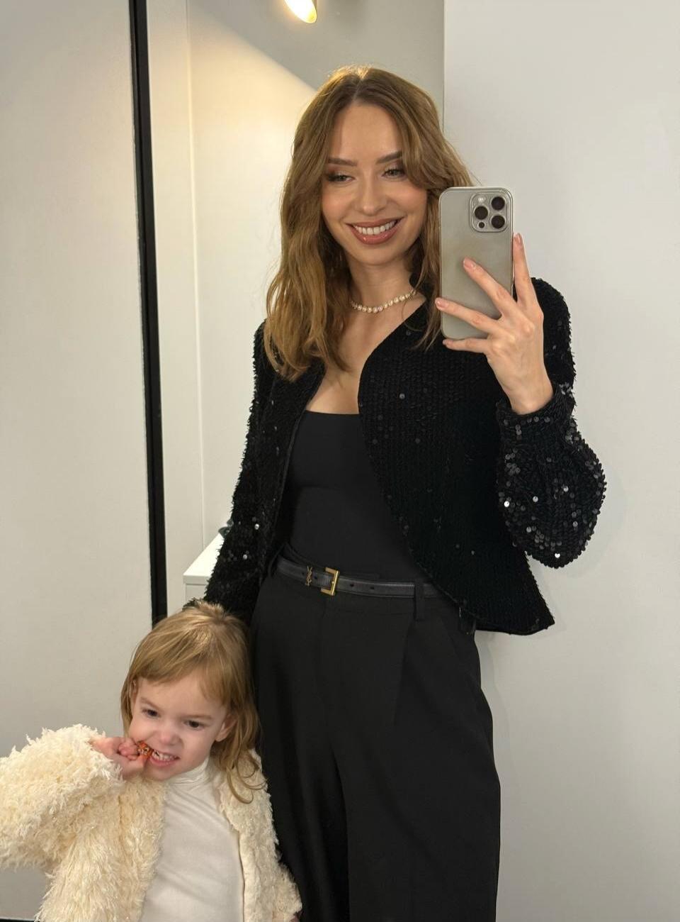 A stylish woman in a black sequined jacket and black outfit takes a mirror selfie with a young child wearing a white fluffy coat.