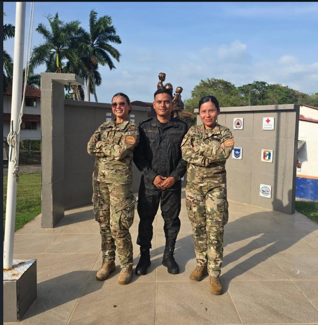 Three individuals in military or police uniforms, two women in camouflage and one man in a dark uniform, stand outdoors. A flag pole is on the left, palm trees are in the background, and a wall with various organizational emblems, including a red cross, is on the right. A sculpture is visible behind the man. Text 'O POS' is on one woman's uniform, 