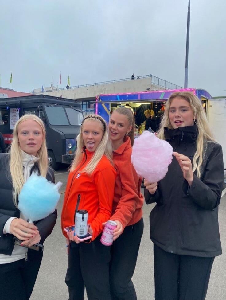 Four young women stand together outdoors, each holding cotton candy. Two wear bright jackets (orange/red), two wear dark outerwear. They appear to be at a fair or event with a colorful trailer in the background.
