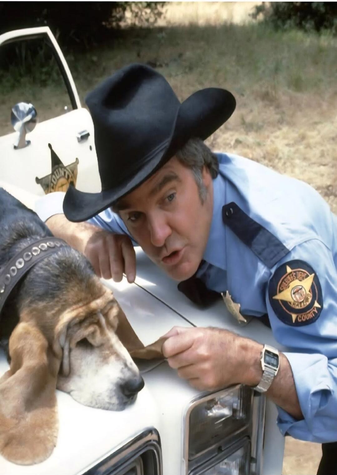 A man in a sheriff's uniform and a cowboy hat leans over a police car, gently interacting with a basset hound. The dog is resting on the car's hood. The sheriff's patch reads 'Hazzard County'.