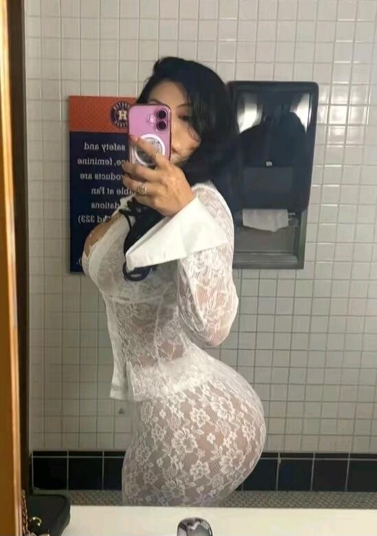 A woman taking a mirror selfie in a restroom, wearing a sheer white lace outfit. A reversed sign is visible in the background, possibly related to safety or animal protection.