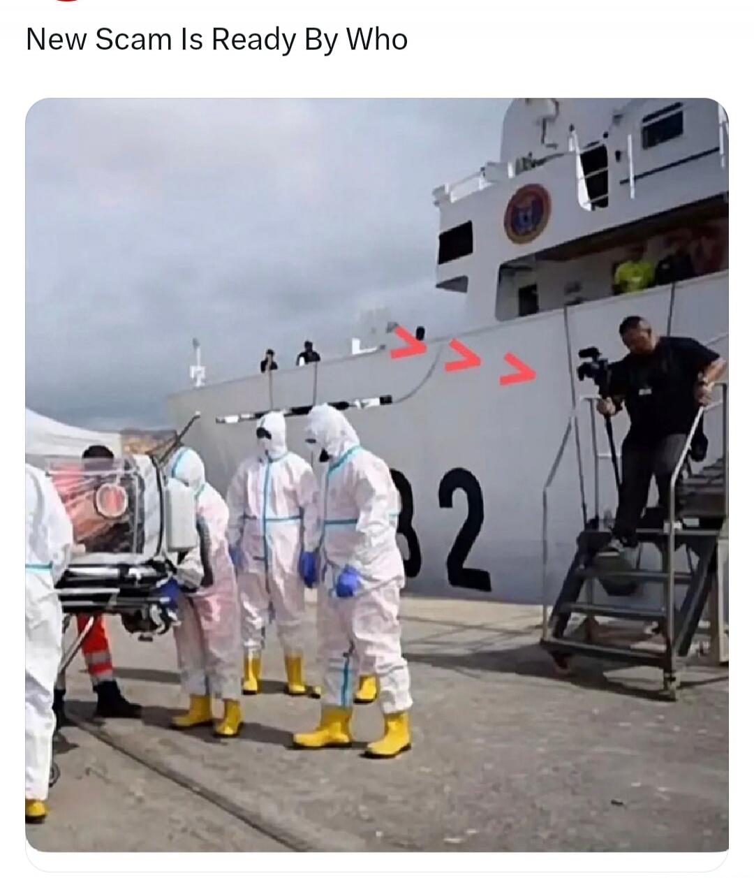 New Scam Is Ready By Who. People in hazmat suits are transferring a patient in an isolation pod onto a ship. A person is filming the scene.