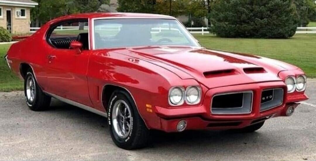 A classic red muscle car, possibly a Pontiac GTO, parked outdoors on an asphalt surface with a house and trees in the background.