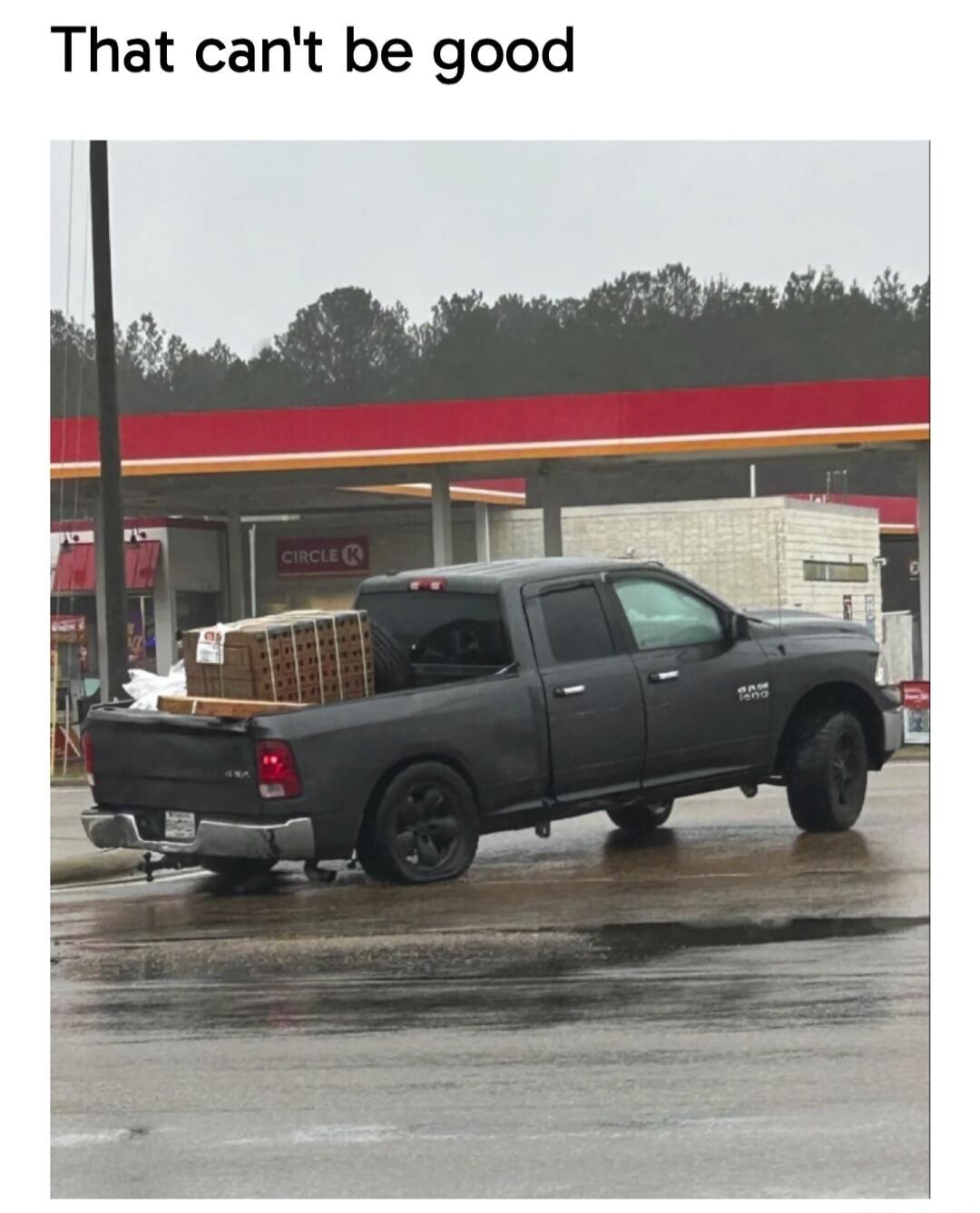 That can't be good. A black pickup truck, a Dodge Ram 1500, is heavily loaded with bricks or pavers in its bed, causing the rear left tire to be completely flat. The truck is parked on a wet road near a Circle K gas station.