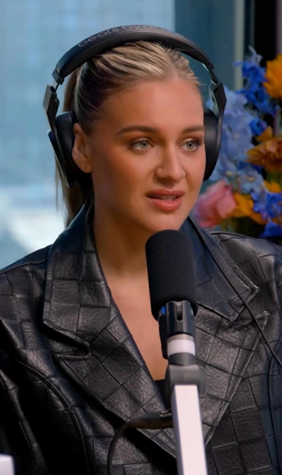 A woman wearing Beats by Dr. Dre headphones and a black quilted leather jacket is speaking into a microphone. She has blonde hair and a focused expression. In the background, out-of-focus flowers are visible.