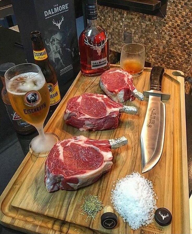 The Dalmore bottle, a boxed The Dalmore display, Eisenbahn beer bottle, a glass of beer, a whiskey bottle and a decanter, three uncooked ribeye steaks with bones on a wooden cutting board, a chef's knife, a pile of coarse salt, a sprig of rosemary, and two small black caps on the cutting board. The scene looks like a meat prep setup with alcohol in