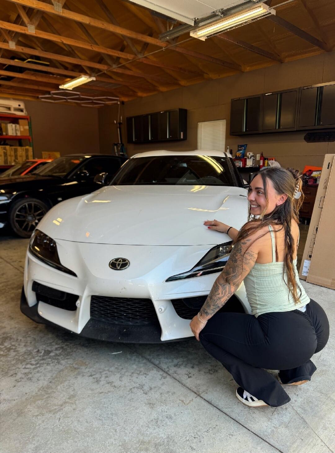 White Toyota sports car in a garage with a smiling woman posing beside the car.