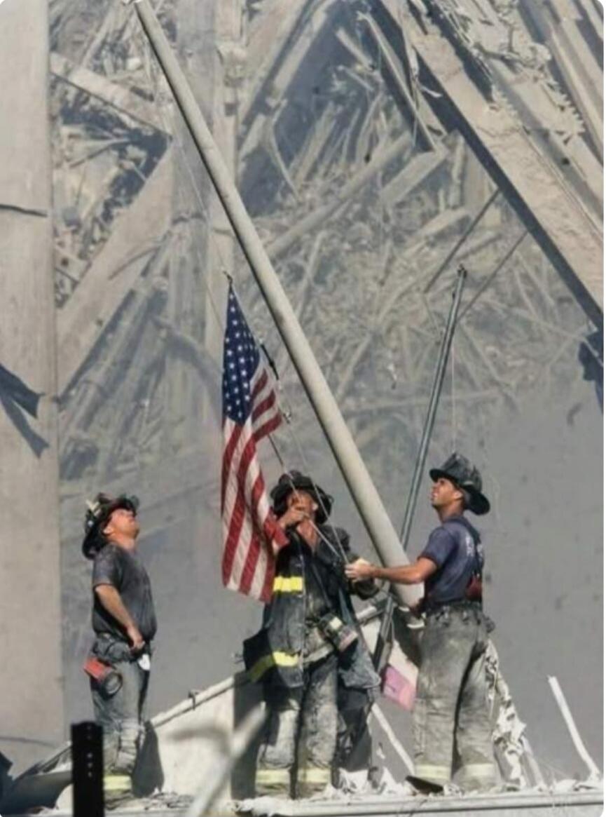 Three firefighters are seen raising an American flag amidst the extensive rubble and dust, likely at Ground Zero after the 9/11 attacks. The scene conveys a powerful message of resilience and patriotism in the face of devastation.