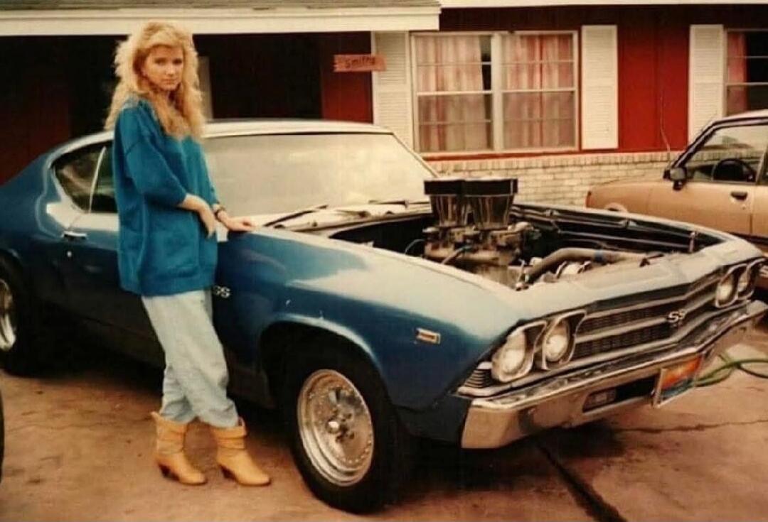 A woman in a blue sweater and light-colored pants stands next to a blue Chevrolet Chevelle SS with its hood open, revealing a powerful engine with dual carburetors. The car appears to be from the late 1960s or early 1970s. The woman has blonde, voluminous hair and is wearing tan boots. The scene is set in front of a house with red siding and white 