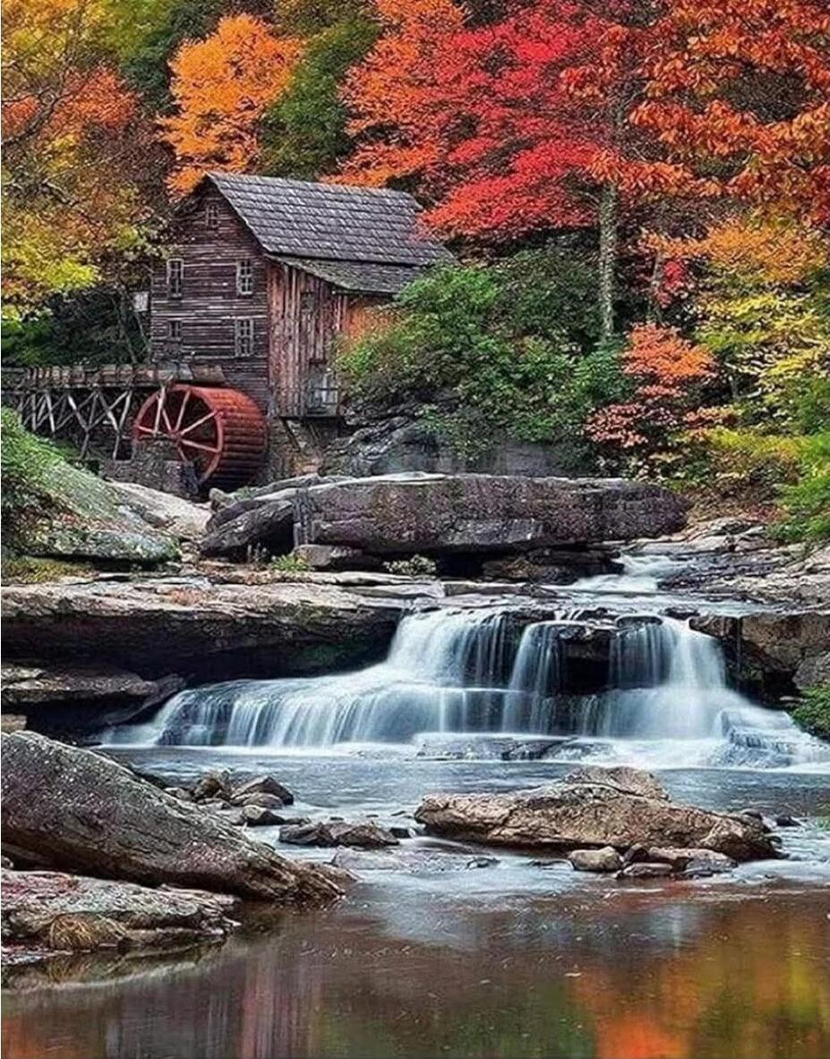 A rustic wooden mill sits beside a small waterfall in a forest, surrounded by vibrant autumn foliage in red, orange, and yellow. A water wheel is attached to the side of the mill, and a gentle stream flows over rocks beneath it.