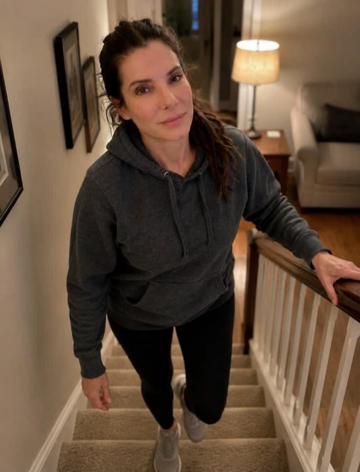 A woman is walking up a carpeted staircase in a home, looking towards the camera with a gentle smile.