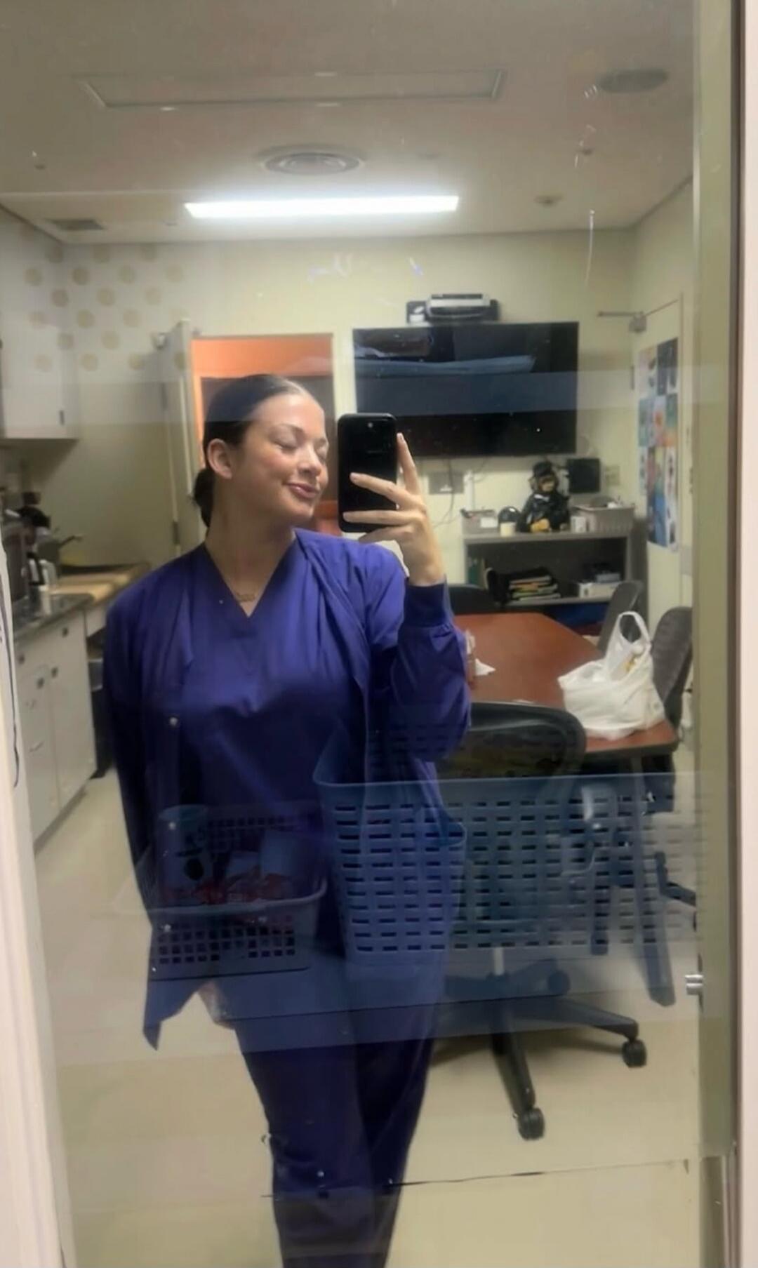 A young woman in blue scrubs takes a mirror selfie, smiling and holding her phone. The reflection shows an office or break room setting.