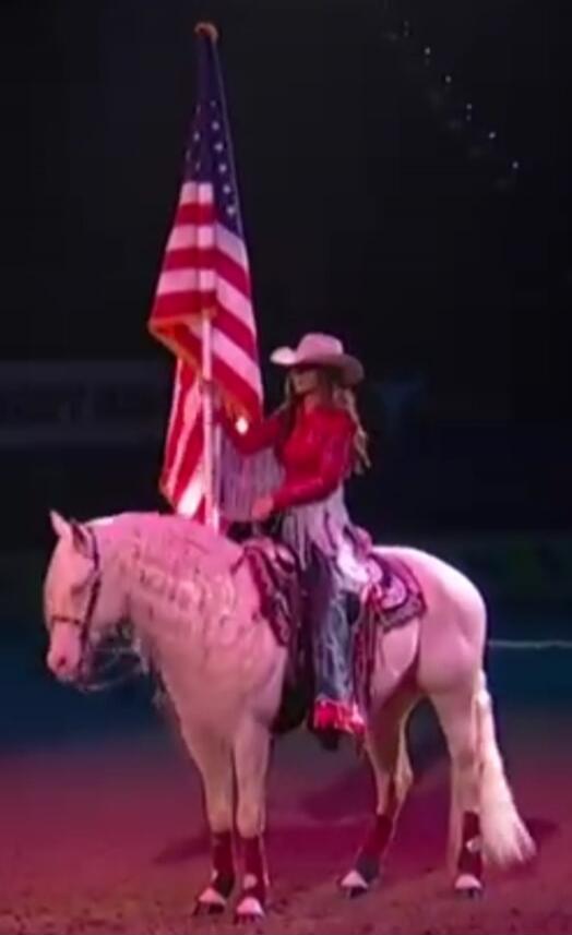 A woman in a cowboy hat and a red sequined shirt rides a white horse, holding an American flag.