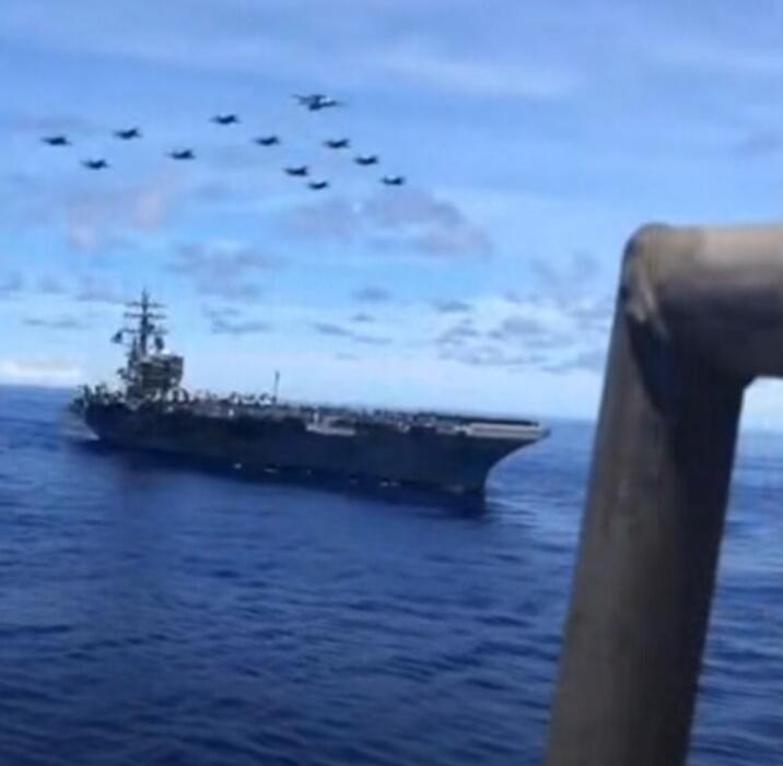An aircraft carrier sails on the open sea with a formation of military aircraft flying overhead in a clear blue sky.