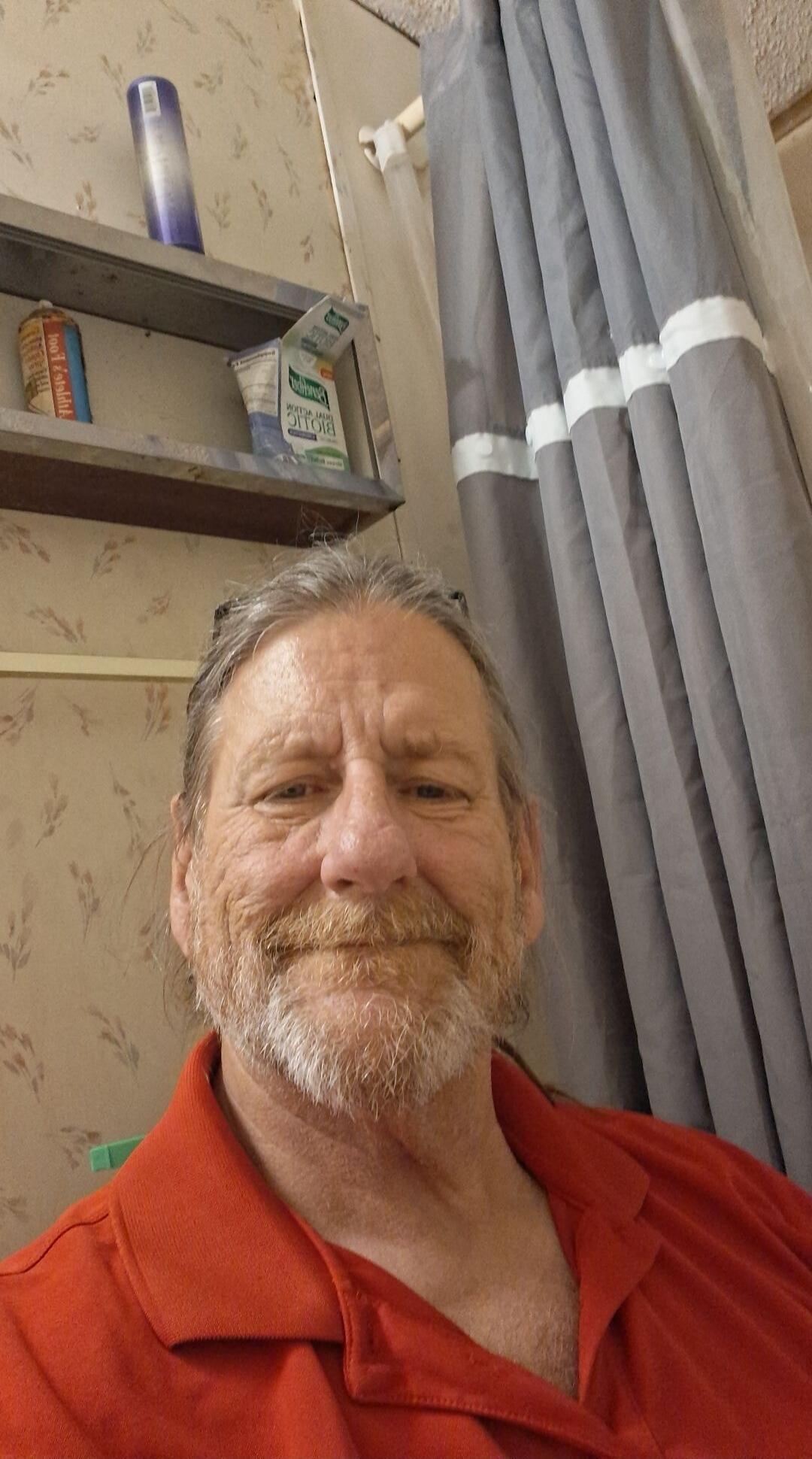 A man with a beard and long hair takes a selfie in a bathroom. He is wearing a red polo shirt. In the background, there is a shower curtain and a shelf with various toiletries.