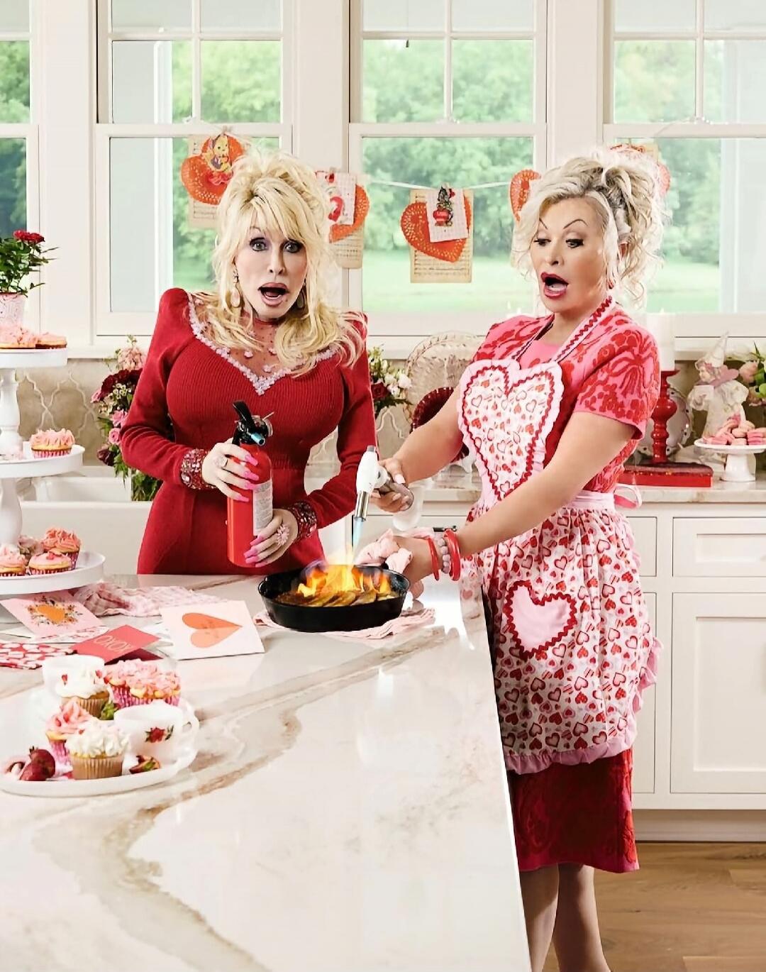 XOXO. Two women, one resembling Dolly Parton, are in a kitchen decorated for Valentine's Day. One woman holds a fire extinguisher, looking surprised, while the other uses a blowtorch on a pan that is on fire. There are cupcakes and heart decorations around.