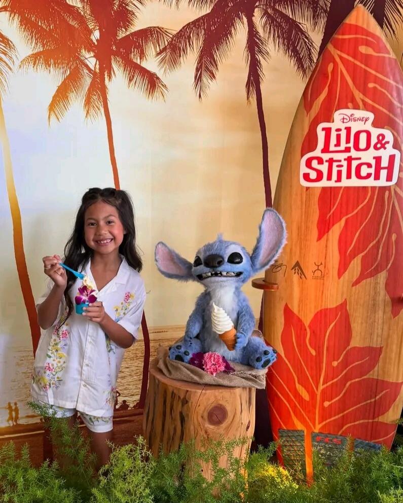 A young girl smiles while holding a cup of ice cream with an orchid, standing next to a Stitch plush toy that also holds a miniature ice cream cone. In the background, there's a tropical scene with palm trees and a surfboard featuring the 'Disney Lilo & Stitch' logo.