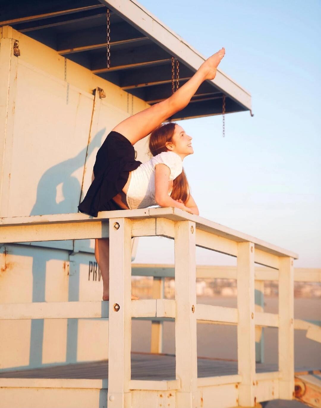 A young woman performing a backbend with one leg extended upwards on a wooden structure, possibly a lifeguard stand, at the beach. Faint text 'PRV' is visible on the structure.