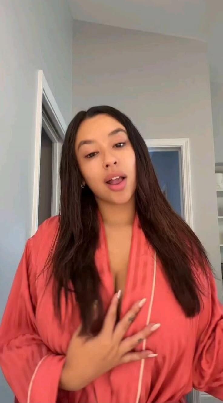 A young woman in a pink robe is speaking to the camera.
