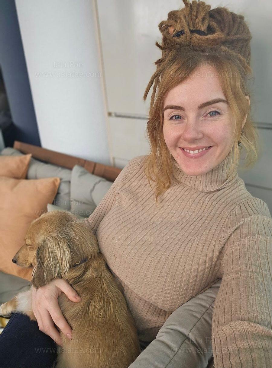 A woman is sitting on a sofa holding a small brown dog, taking a selfie.