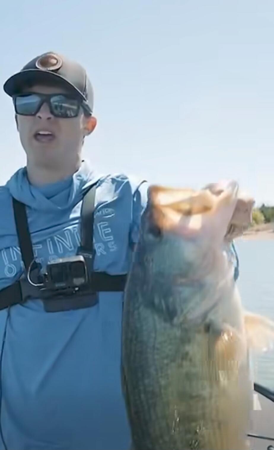 A man wearing a blue shirt, sunglasses, and a GoPro on a chest strap is holding a large fish on a boat.
