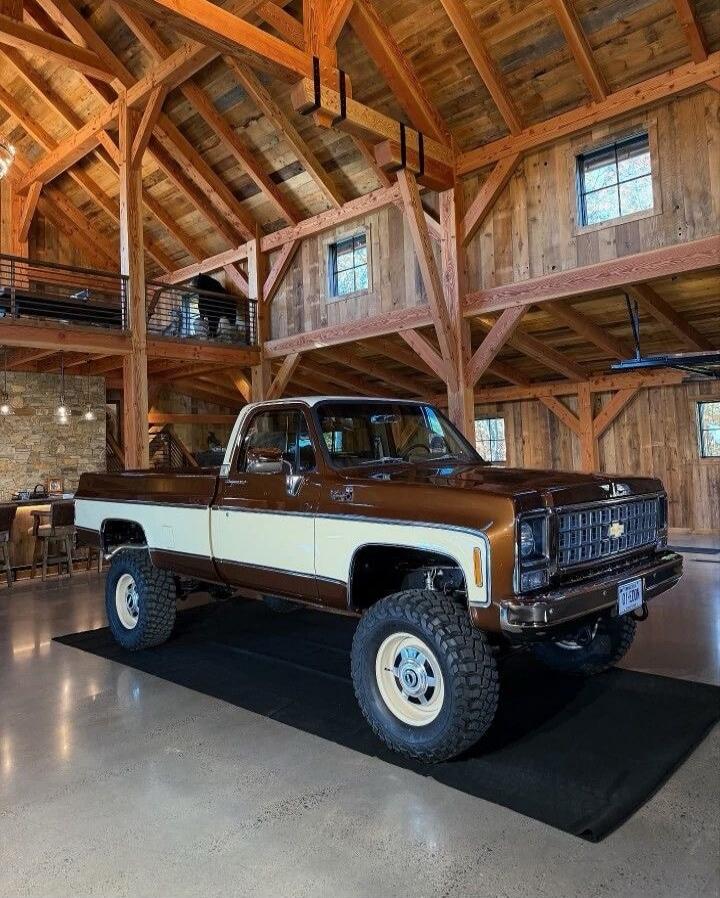 A vintage brown and white pickup truck is displayed inside a spacious wooden barn-like showroom with exposed beams and high ceilings. The truck has large off-road tires and sits on a dark rug. The background shows wooden walls, windows, and a mezzanine area.