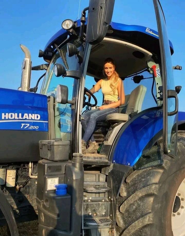 HOLLAND T7.200 Bluepower. A young woman is sitting in the driver's seat of a large blue tractor, smiling at the camera.