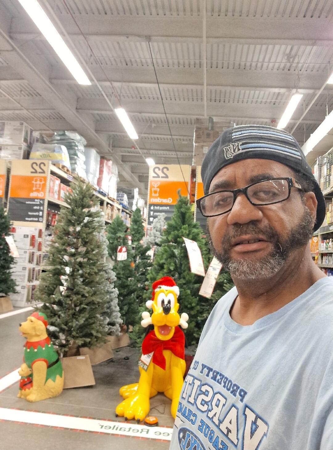 A man takes a selfie in a store aisle filled with artificial Christmas trees and holiday decorations. In the background, there are large figures of Pluto wearing a Santa hat and a dog in an elf costume. The man is wearing a t-shirt with 'PROPERTY OF VARSITY ATHLETIC DEPT.' printed on it (reversed) and a cap. Store signs like 'SS' and 'REGISTER 99' 