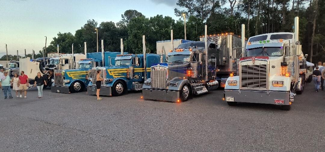 A line of custom semi-trucks with chrome details and unique paint jobs parked outdoors, with people walking around and admiring them.